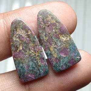Peut inclure: Deux pierres précieuses polies avec un mélange de couleurs. Les pierres ont un dessus arrondi et un fond plat. Les couleurs comprennent le rouge profond, le sarcelle, l'or et le gris. Les pierres précieuses sont probablement utilisées pour la fabrication de bijoux ou la collection.