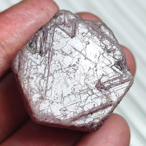 May include: A raw, hexagonal-shaped gemstone with a pale pinkish-purple hue. The surface displays a complex network of intersecting linear fractures and crystalline striations, creating a textured, reflective appearance across the flat face.