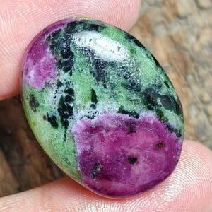 May include: An oval-shaped polished gemstone featuring a vibrant mix of deep magenta-pink ruby inclusions set within a light green zoisite matrix, accented by scattered dark black hornblende mineral deposits across the smooth, reflective surface.