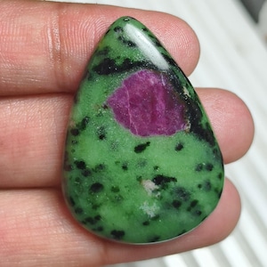 Puede incluir: Una piedra preciosa de zoisita pulida en forma de lágrima, con una base de color verde vibrante, inclusiones minerales negras dispersas y una formación central prominente de cristal de rubí magenta opaco con una superficie lisa y brillante.