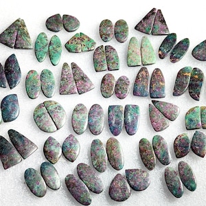 Könnte beinhalten: Eine Auswahl an polierten Edelsteinen in verschiedenen Formen, darunter Ovale, Dreiecke und Tropfen. Die Steine zeigen eine Mischung aus Farben, mit dominanten Grüntönen, Lila und goldenen Akzenten. Die Edelsteine sind wahrscheinlich für die Schmuckherstellung oder das Basteln gedacht.