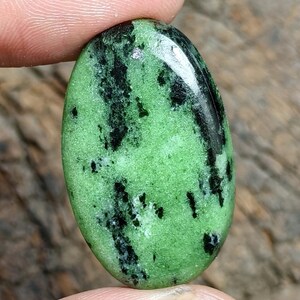 Può includere: Una pietra preziosa a cabochon di forma ovale caratterizzata da un colore di base verde lime vibrante con evidenti inclusioni minerali nere irregolari distribuite sulla superficie lucida. La pietra presenta una finitura liscia e bombata.