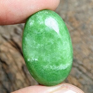 Può includere: Una gemma verde lucida di forma ovale con una superficie liscia a cabochon. La pietra presenta un colore verde vibrante e traslucido con sottili inclusioni minerali bianche e texture interne naturali visibili sulla superficie lucida.