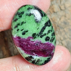 Puede incluir: Una piedra preciosa cabujón de forma ovalada que presenta una base de color verde vibrante con inclusiones minerales negras dispersas y una banda horizontal texturizada prominente de cristal color magenta intenso en la parte inferior.