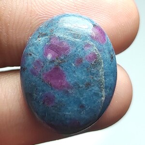 Op de afbeelding: Ovale edelsteen met een turquoise basiskleur en levendige magenta insluitsels. De gepolijste steen heeft een glad oppervlak en een natuurlijke uitstraling. De edelsteen wordt tussen twee vingers gehouden. De steen is ongeveer 2,5 cm lang.