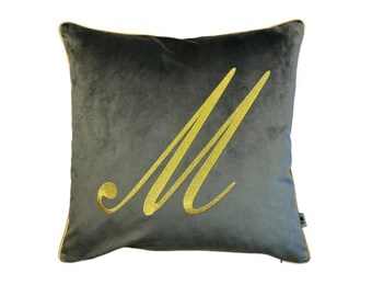 May include: A gray velvet pillow with a gold embroidered letter "M" in a cursive font.