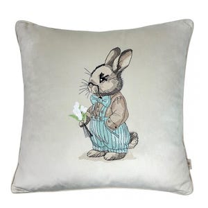 May include: A light gray throw pillow with an embroidered image of a brown rabbit wearing a blue bow tie and blue overalls holding a bouquet of white flowers.