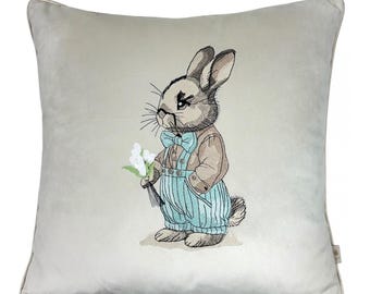 Könnte beinhalten: Ein hellgraues Kissen mit einem gestickten Bild eines braunen Kaninchens, das eine blaue Fliege und eine blaue Latzhose trägt und einen Strauß weißer Blumen hält.