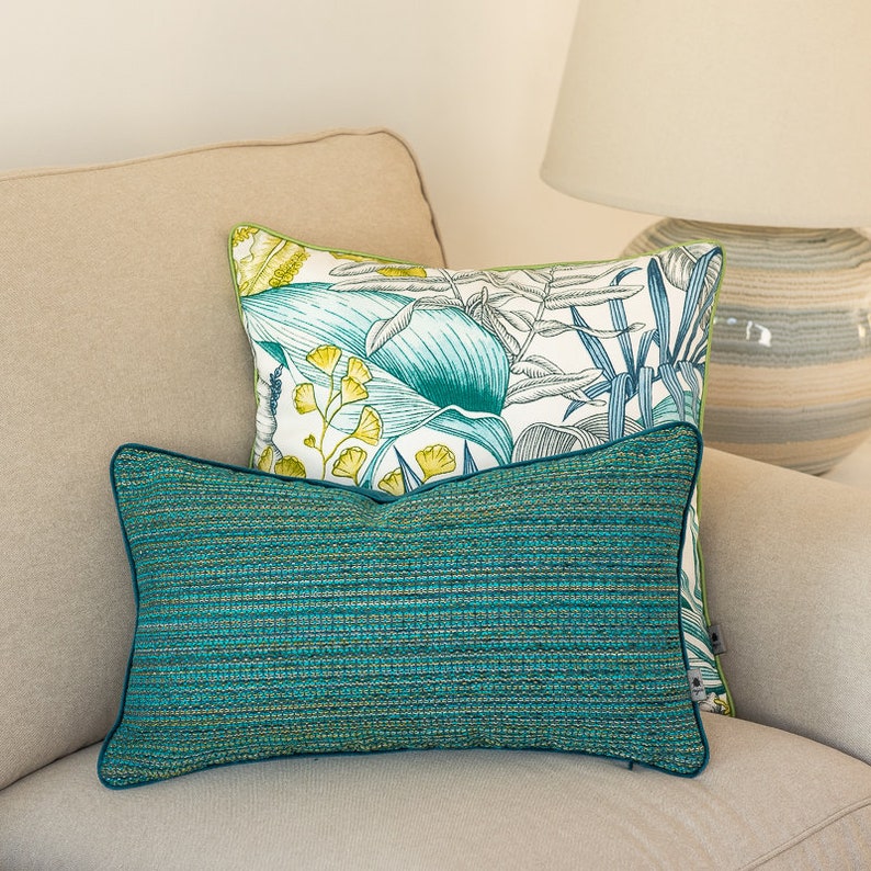 K&ouml;nnte beinhalten: Zwei dekorative Kissen auf einem beigen Sofa. Das obere Kissen hat einen wei&szlig;en Hintergrund mit einem tropischen Blattmotiv in Gr&uuml;n und Blau. Das untere Kissen ist einfarbig in Teal mit einer dezenten gewebten Textur.