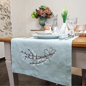 Könnte beinhalten: Ein hellblauer Tischläufer aus Leinen mit einem gestickten Muster aus braunen Weidenzweigen mit weißen Blüten. Der Läufer liegt auf einem Holztisch mit einer weißen Tischdecke und einem Gedeck.