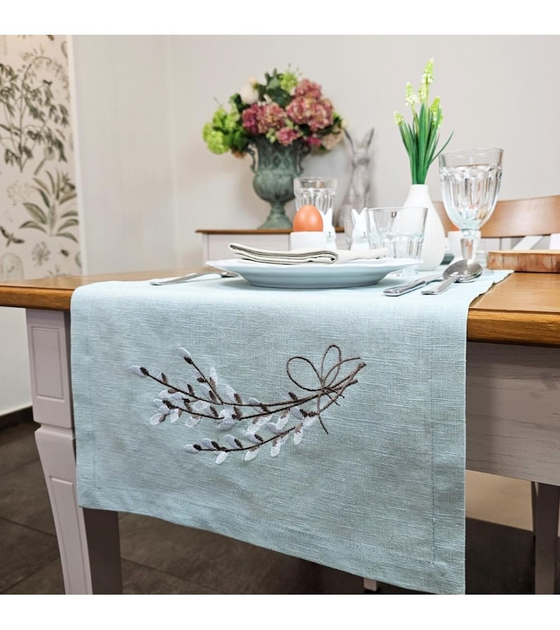 Puede incluir: Un camino de mesa de lino azul claro con un dise&ntilde;o bordado de ramas de sauce marrones con flores blancas. El camino de mesa est&aacute; colocado sobre una mesa de madera con un mantel blanco y una vajilla.