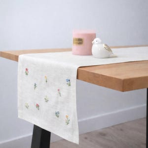 Puede incluir: Camino de mesa crema con flores coloridas bordadas. Una vela rosa y una figura de pájaro blanco con detalles dorados están sobre la mesa de madera. La mesa tiene una pata negra.