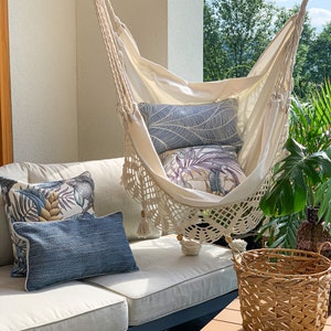 May include: A white hammock with crocheted trim hangs from the ceiling of a patio. The hammock is decorated with pillows featuring tropical leaf patterns. A wicker basket sits on a wooden table in the foreground.
