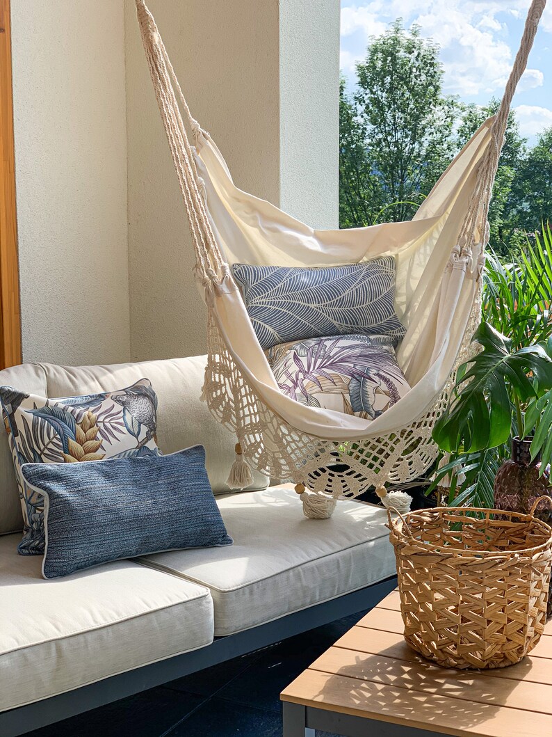 May include: A white hammock with crocheted trim hangs from the ceiling of a patio. The hammock is decorated with pillows featuring tropical leaf patterns. A wicker basket sits on a wooden table in the foreground.