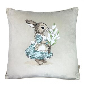 May include: A beige velvet pillow with an embroidered image of a rabbit wearing a blue dress and holding a bouquet of white flowers.