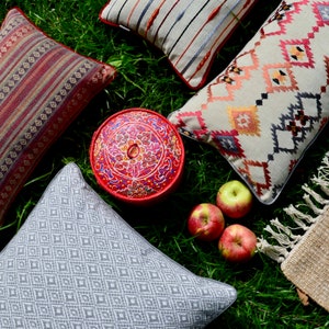 May include: A collection of decorative throw pillows with various patterns and colors, including red, white, and blue. The pillows are arranged on a bed of green grass with a red and white painted wooden box in the center. There are also three red apples on the grass.