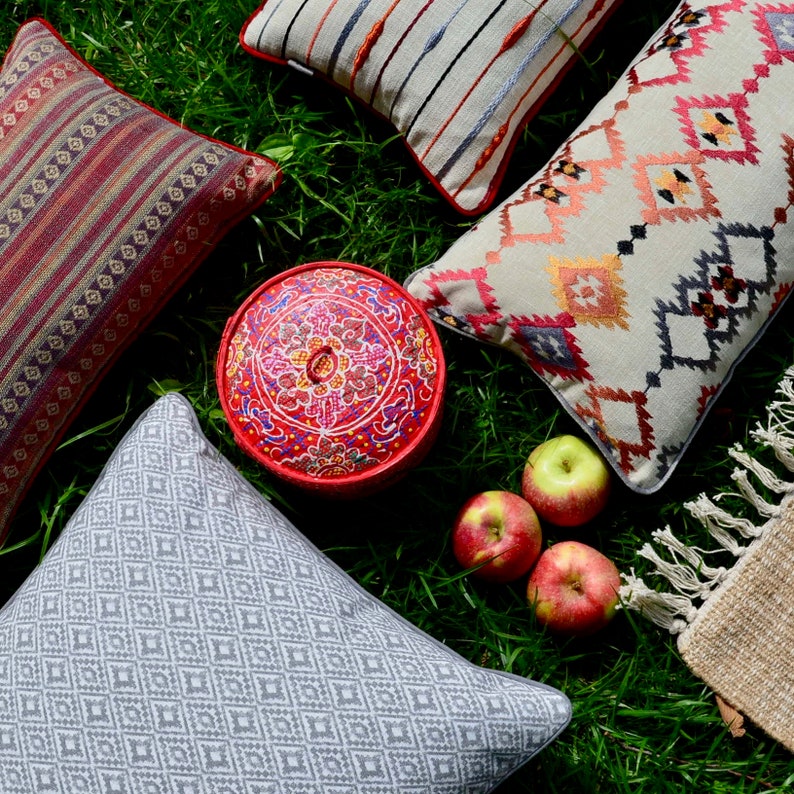 May include: A collection of decorative throw pillows with various patterns and colors, including red, white, and blue. The pillows are arranged on a bed of green grass with a red and white painted wooden box in the center. There are also three red apples on the grass.