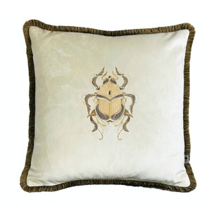May include: A cream-colored velvet pillow with a brown fringe border features an embroidered gold and gray beetle design.