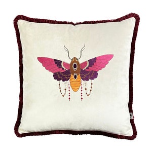 Könnte beinhalten: Ein weißes Samtkissen mit einem burgunderfarbenen Fransenrand zeigt eine gestickte Motte mit rosa, violetten und orangefarbenen Akzenten. Die Motte hat eine Perlenkette, die von ihrem Körper hängt.