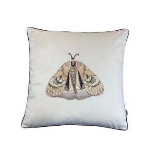May include: A square, light gray decorative pillow with a detailed embroidered moth design in shades of brown, tan, and black. The pillow has a black trim around the edges, adding a contrasting border to the design.