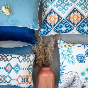 Pu&ograve; includere: Una collezione di cuscini decorativi con vari motivi e colori. I cuscini presentano disegni geometrici, motivi floreali e un motivo ikat blu e bianco. I cuscini sono disposti su una superficie grigio chiaro con un vaso di erba secca al centro.
