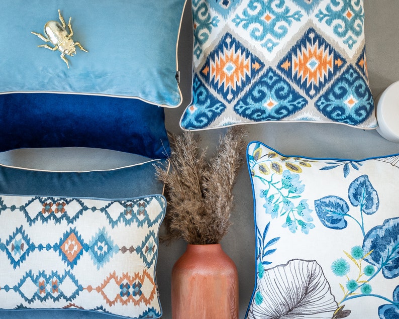 Pu&ograve; includere: Una collezione di cuscini decorativi con vari motivi e colori. I cuscini presentano disegni geometrici, motivi floreali e un motivo ikat blu e bianco. I cuscini sono disposti su una superficie grigio chiaro con un vaso di erba secca al centro.