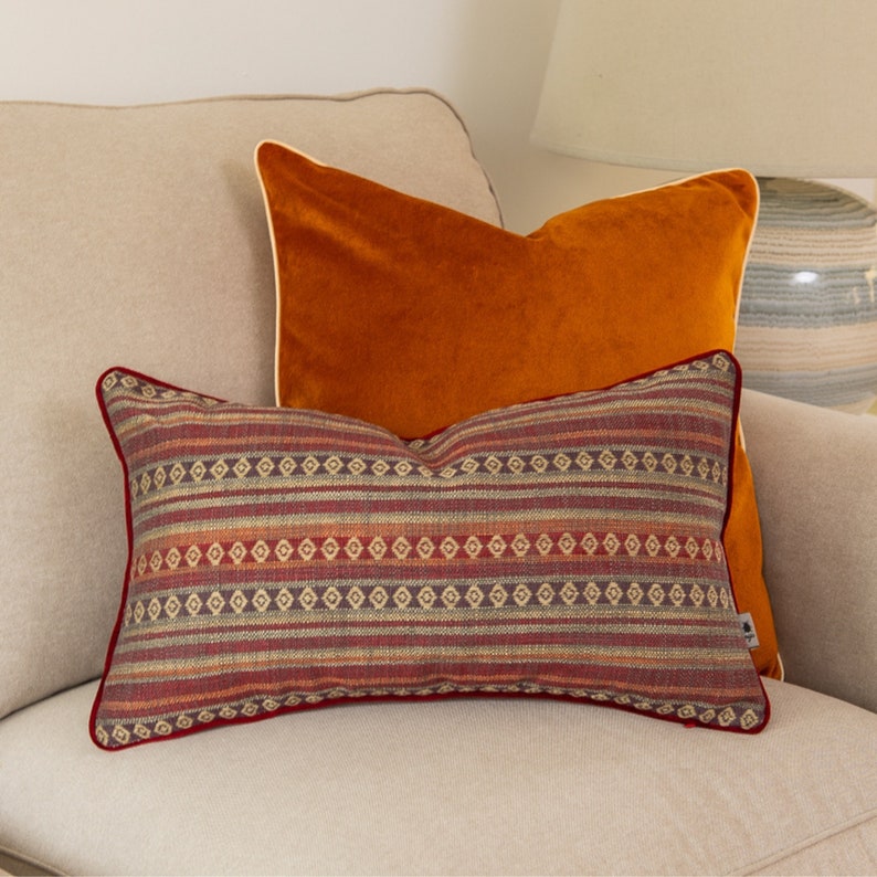 May include: Two decorative throw pillows, one with a geometric pattern in shades of red, orange, and brown, and the other a solid orange velvet pillow with a cream trim.
