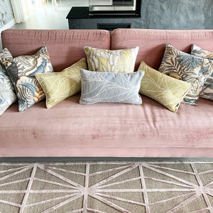 May include: A pink velvet sofa with a variety of throw pillows featuring tropical leaf patterns. The pillows are in shades of green, blue, and white. The sofa is sitting on a gray and white geometric patterned rug.