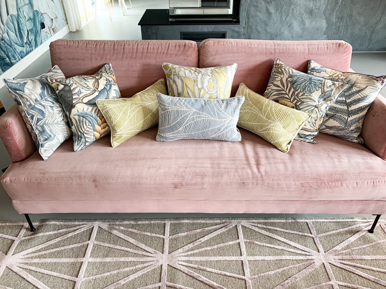 May include: A pink velvet sofa with a variety of throw pillows featuring tropical leaf patterns. The pillows are in shades of green, blue, and white. The sofa is sitting on a gray and white geometric patterned rug.