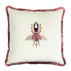 Könnte beinhalten: Ein weißes Kissen mit einem burgunderfarbenen Fransenrand zeigt ein gesticktes Design eines Hirschkäfers mit Flügeln, einem Halbmond und einem Stern.