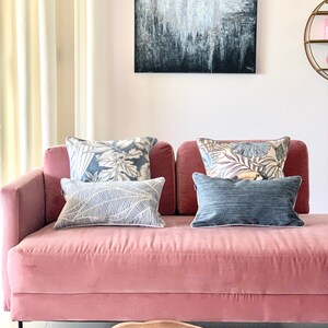 May include: A pink velvet sofa with a black metal frame and four throw pillows. Two of the pillows have a floral pattern, one has a leaf pattern, and one is solid blue. The sofa is in front of a white wall with a large abstract painting above it.