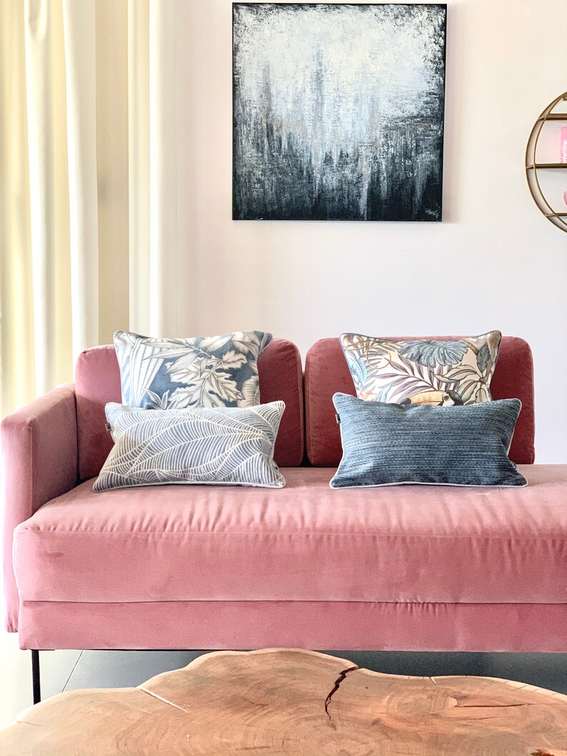May include: A pink velvet sofa with a black metal frame and four throw pillows. Two of the pillows have a floral pattern, one has a leaf pattern, and one is solid blue. The sofa is in front of a white wall with a large abstract painting above it.