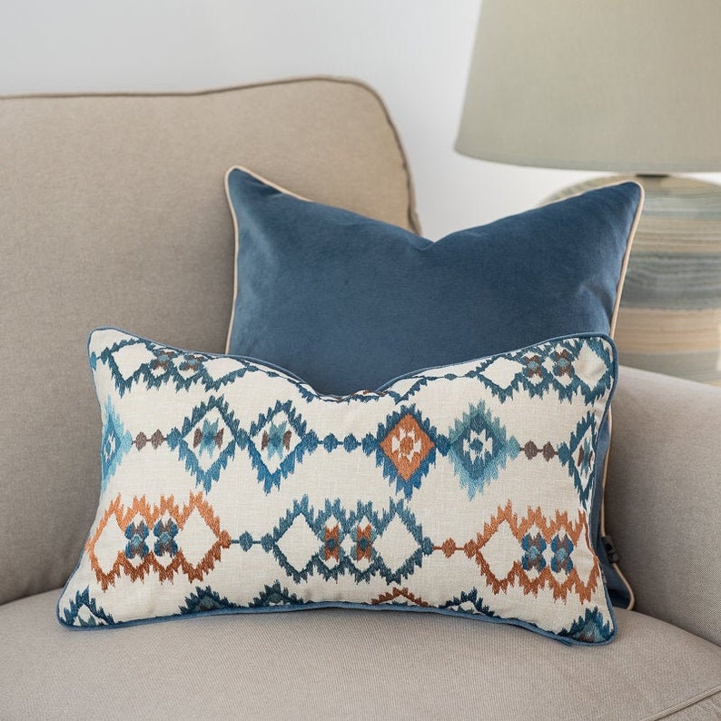 Pu&ograve; includere: Due cuscini decorativi su un divano beige. Il cuscino inferiore presenta un motivo geometrico in tonalit&agrave; di blu, arancione e bianco. Il cuscino superiore &egrave; in velluto blu tinta unita.