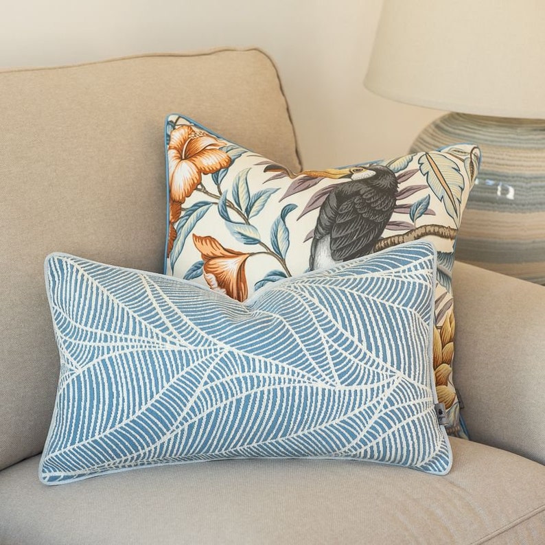 May include: Two decorative throw pillows on a beige couch. The front pillow is a light blue with a white leaf pattern. The back pillow is white with a tropical floral pattern and a toucan.