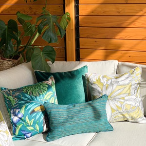 K&ouml;nnte beinhalten: Ein wei&szlig;es Ecksofa mit mehreren Kissen. Die Kissen sind gr&uuml;n, gelb und blau mit tropischen Mustern. Das Sofa steht in einem Raum mit einer Holzwand und einer gr&uuml;nen Pflanze.