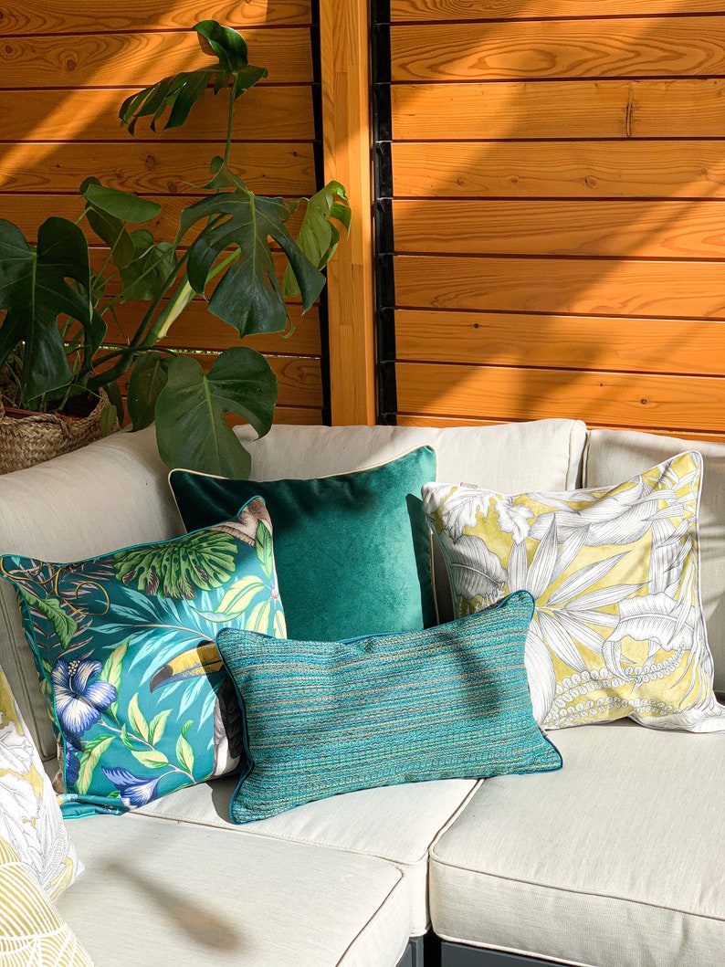 K&ouml;nnte beinhalten: Ein wei&szlig;es Ecksofa mit mehreren Kissen. Die Kissen sind gr&uuml;n, gelb und blau mit tropischen Mustern. Das Sofa steht in einem Raum mit einer Holzwand und einer gr&uuml;nen Pflanze.
