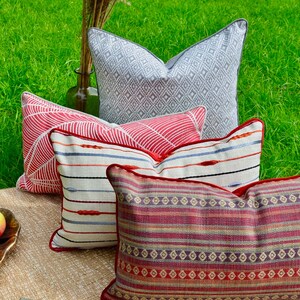 May include: Four decorative throw pillows with different patterns. The pillows are arranged in a stack with the top pillow being a gray and white geometric pattern, the second pillow is a red and white geometric pattern, the third pillow is a cream and blue striped pattern, and the bottom pillow is a red, cream, and purple striped pattern.