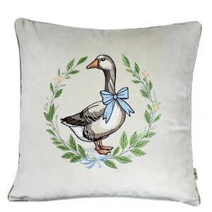 May include: A white velvet pillow with an embroidered design of a goose wearing a blue bow. The goose is surrounded by a wreath of green leaves and small white flowers.