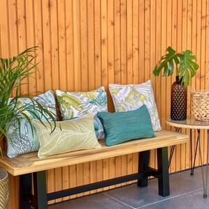K&ouml;nnte beinhalten: Eine Holzbank mit schwarzen Metallbeinen, ausgestattet mit vier dekorativen Kissen mit tropischen Blattmotiven. Die Bank steht auf einer Terrasse mit einer Holzwand im Hintergrund.