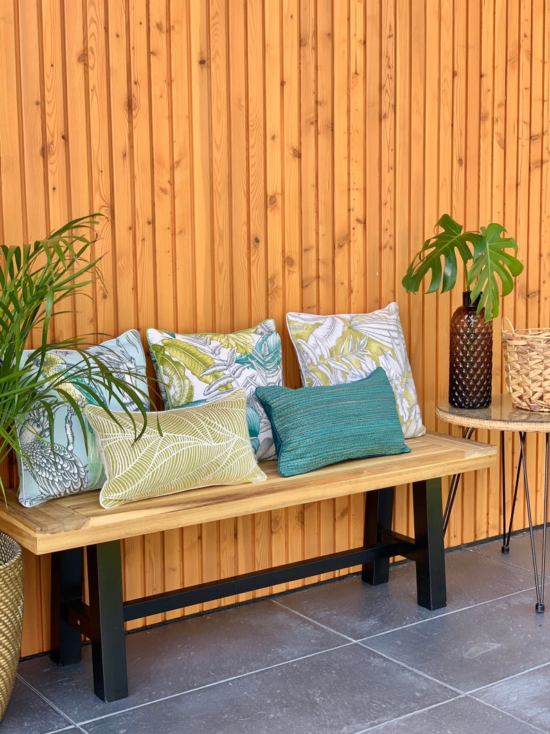 K&ouml;nnte beinhalten: Eine Holzbank mit schwarzen Metallbeinen, ausgestattet mit vier dekorativen Kissen mit tropischen Blattmotiven. Die Bank steht auf einer Terrasse mit einer Holzwand im Hintergrund.
