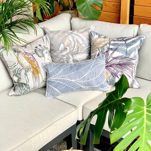May include: A white couch with four throw pillows featuring tropical leaf and bird patterns. The couch is set against a wooden wall with a potted plant in the background.