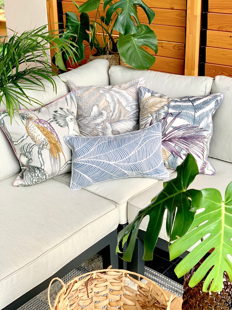 May include: A white couch with four throw pillows featuring tropical leaf and bird patterns. The couch is set against a wooden wall with a potted plant in the background.
