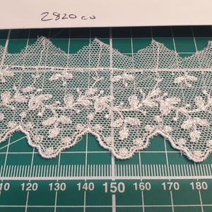 Puede incluir: Ribete de encaje color marfil con borde festoneado y diseño floral. El encaje está sobre un fondo de cuadrícula verde con medidas en centímetros. El texto "2820 cu" e "Ivory" son visibles.