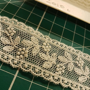May include: Close-up of a cream-colored lace trim with a floral design. The lace has a delicate, openwork pattern with a scalloped edge. The text "2010c" is written on the backing.