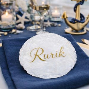 Può includere: Un'impostazione della tavola a tema nautico con accenti dorati. Un piatto bianco con un tovagliolo blu navy e un caricatore dorato. Un segnaposto bianco con il nome "Rurik" in oro. Decorazioni e posate a forma di ancora dorata.