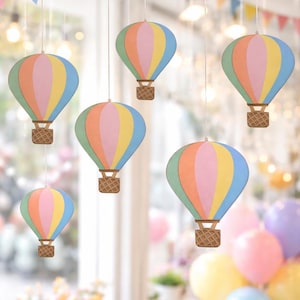 May include: Pastel-coloured hot air balloon decorations suspended from the ceiling. Each balloon has pink, yellow, blue, and green panels. The balloons have a woven basket design at the bottom, and are hung by thin white strings. Party decorations.