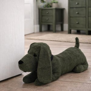 May include: A dark olive green fabric dog doorstop with long floppy ears, a black nose, and a black collar. The plush dog is in a lying position with its tail pointed upward, featuring a textured woven material finish throughout its body.