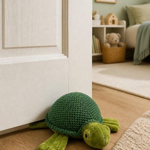 May include: A turtle-shaped doorstop featuring a dark green crocheted shell and a light green plush body with four flippers and a head with black eyes. The turtle is positioned on a wooden floor, wedged against the base of a white door.