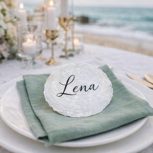 Peut inclure: Une carte de place blanche et texturée avec le nom "Lena" en écriture noire élégante est posée sur une serviette en lin vert sauge. L'ensemble comprend une assiette blanche, des couverts dorés et une plage en arrière-plan.