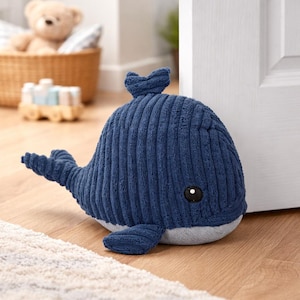 Peut inclure: Un butoir de porte en forme de baleine bleu marine en tissu côtelé. Le butoir de porte a un ventre blanc, une petite nageoire et une queue. Il a un œil noir et repose sur un tapis clair et un sol en bois.